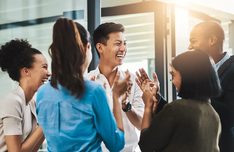 Un groupe de six personnes se tient dans un bureau, riant et discutant joyeusement ensemble. Elles semblent célébrer un moment de convivialité et de réussite, avec des expressions heureuses et des gestes d'approbation. La lumière du jour entre par des fenêtres.