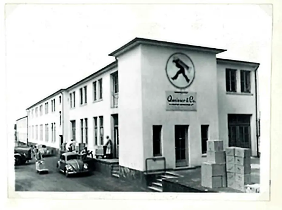 Un bâtiment en noir et blanc avec une enseigne circulaire représentant un personnage en marche. Des hommes et femmes sont visibles à l'extérieur, à côté de caisses empilées. Des voitures anciennes sont garées devant l'entrée de l'immeuble.
