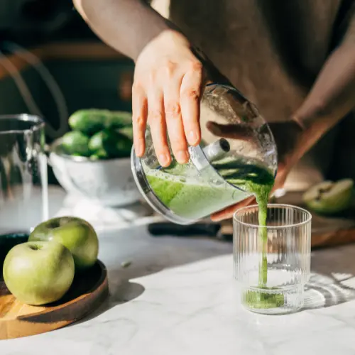 Une main verse un smoothie vert dans un verre transparent. Sur la table, on peut voir des pommes vertes et un bol de concombres, évoquant une préparation saine. La lumière du jour illumine la scène, mettant en valeur les textures des ingrédients.