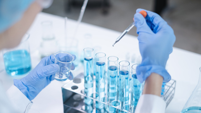 Un scientifique en blouse blanche et gants bleus réalise une expérience en laboratoire. Il tient un bécher rempli d'un liquide et utilise une pipette pour ajouter une goutte dans des tubes à essai contenant un liquide bleu.