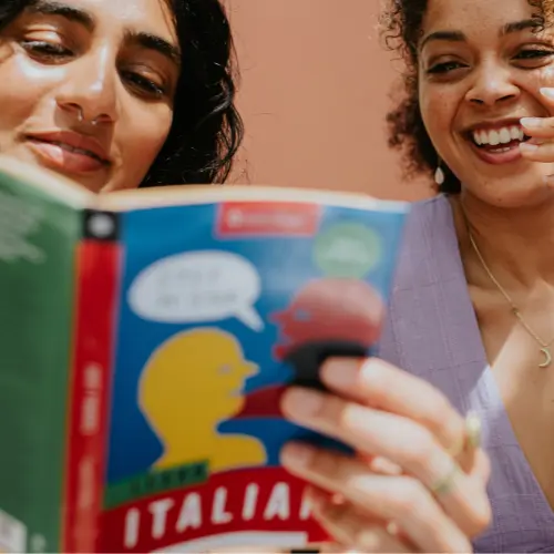 Deux femmes souriantes lisent ensemble un livre sur la langue italienne. L'une tient le livre ouvert, tandis que l'autre regarde avec intérêt, visiblement amusée. Un fond coloré met en valeur leur convivialité et leur enthousiasme pour l'apprentissage.