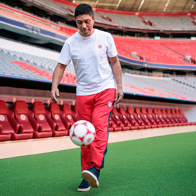 Un homme souriant dribble un ballon de football sur le terrain d'un stade. Il porte un t-shirt blanc et un pantalon rouge avec des détails bleus. En arrière-plan, on aperçoit des sièges rouges dans le stade.