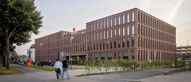 Un bâtiment moderne en briques avec de grandes fenêtres, situé en milieu urbain. Des passants marchent sur un trottoir, avec des voitures garées à proximité. Un logo rouge est visible sur le toit. Des arbres et des buissons encadrent le site.