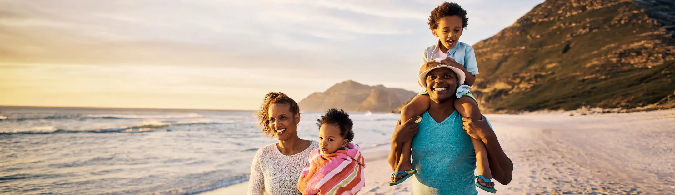 Quatre personnes se tiennent sur la plage, avec l'océan en arrière-plan et des montagnes. Un homme porte un enfant sur ses épaules, tandis qu'une femme tient un bébé enroulé dans une serviette. Tous affichent des sourires joyeux sous un ciel ensoleillé.
