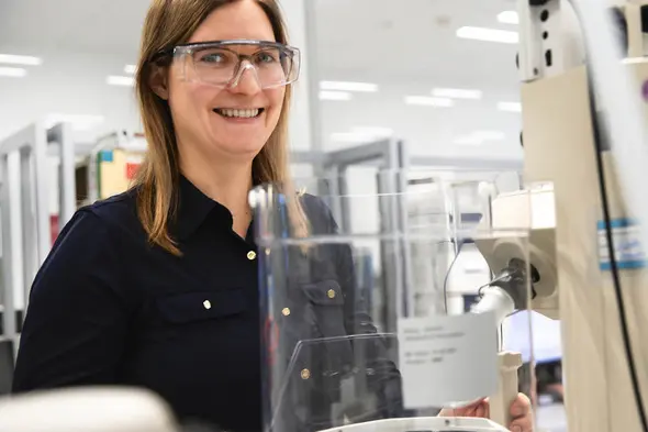 Une femme souriante portant des lunettes de protection se tient devant un équipement de laboratoire. Elle semble travailler dans un environnement scientifique moderne, avec des instruments et des ustensiles visibles autour d'elle.