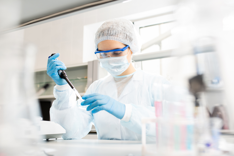 Une scientifique en blouse et portant des gants effectue des mesures avec une pipette dans un laboratoire. Elle porte également un masque et des lunettes de protection. Des instruments et des récipients de laboratoire sont visibles en arrière-plan.