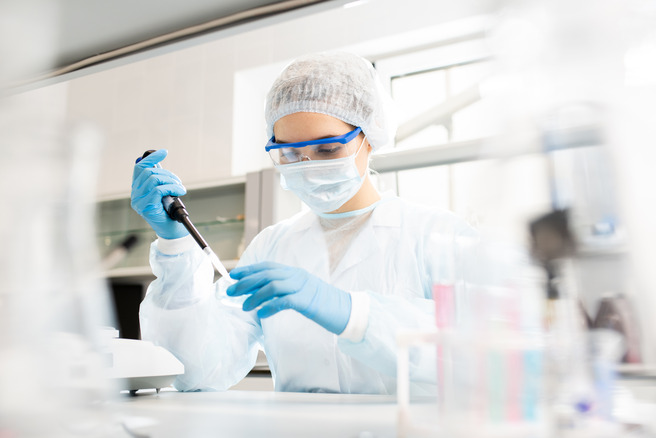 Une scientifique en blouse et portant des gants effectue des mesures avec une pipette dans un laboratoire. Elle porte également un masque et des lunettes de protection. Des instruments et des récipients de laboratoire sont visibles en arrière-plan.