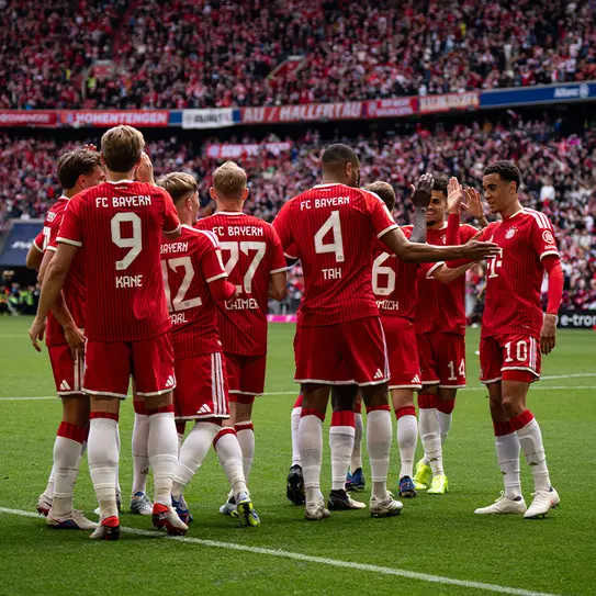 Des joueurs du FC Bayern Munich se regroupent sur le terrain, vêtus de maillots rouges. L'ambiance est festive avec des supporters en arrière-plan. Les joueurs échangent des gestes amicaux avant le début du match.