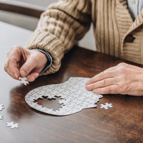 Une main âgée tient une pièce de puzzle, tandis que l'autre main place une pièce dans un puzzle en cours de réalisation sur une table en bois. Le puzzle représente une forme partielle, indiquant un moment de concentration et de détente.