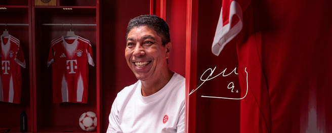 Un homme souriant assis à l'intérieur d'un vestiaire, devant des maillots rouges de l'équipe de football Bayern Munich. Des éléments sportifs comme un ballon de football sont visibles à proximité. L'ensemble de l'image est dominé par des tons rouges.