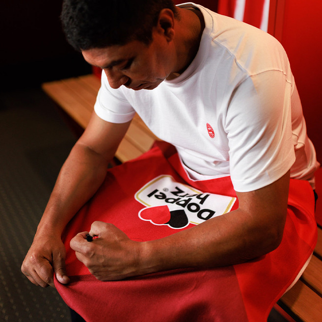 Un homme assis sur un banc dans un vestiaire, portant un t-shirt blanc. Il est en train de soigner un vêtement rouge avec un logo "Popped It". L'ambiance est sportive et décontractée.