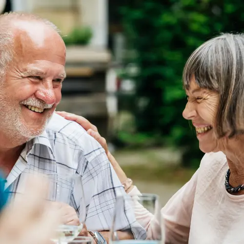 Deux personnes âgées, un homme et une femme, rient ensemble lors d'un repas en plein air. Ils se regardent avec tendresse et complicité, partageant un moment convivial et joyeux. Des verres et des plats sont visibles sur la table à proximité.