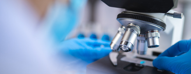 Un laboratoire avec un microscope au premier plan. Des mains en gants bleus ajustent un échantillon sous l'objectif du microscope. L'arrière-plan montre une ambiance de travail scientifique.