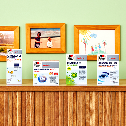 Des boîtes de produits de santé sont alignées sur une table en bois. Les produits incluent "Omega-3 Plus", "Magnésium 400 Citrate", et "Omega-3 Pflanzlich" ainsi que "Augen Plus". En arrière-plan, des photos d'enfants et un dessin ornent un mur de couleur verte.