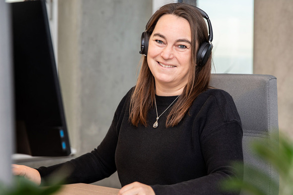 Femme souriante assise à un bureau avec un casque audio. Elle porte un pull noir et se trouve devant un ordinateur. En arrière-plan, on aperçoit des murs en béton et des plantes.