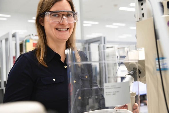 Une femme souriante portant des lunettes de protection se tient devant un équipement de laboratoire. Elle semble travailler dans un environnement scientifique moderne, avec des instruments et des ustensiles visibles autour d'elle.