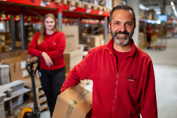 Deux employés dans un entrepôt. Un homme souriant, portant un vêtement rouge, tient une boîte. En arrière-plan, une femme en rouge se tient près d'un transpalette, surveillant l'environnement de travail. Des rangées de boîtes sont visibles autour d'eux.