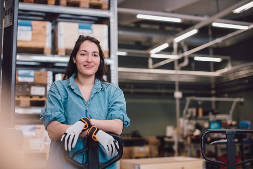Une femme souriante, portant une chemise en jean et des gants de travail, se tient avec les bras croisés devant un chariot élévateur dans un entrepôt. En arrière-plan, des étagères remplies de caisses en bois sont visibles.
