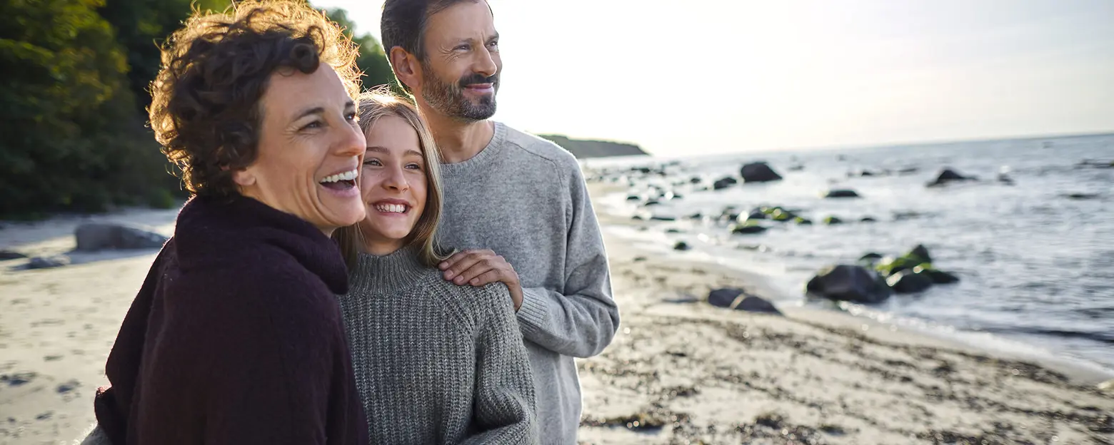 Trois personnes souriantes se tiennent sur une plage, entourées d'un paysage côtier. Deux femmes, l'une aux cheveux bouclés et l'autre avec des cheveux blonds, sont aux côtés d'un homme, tous habillés en pulls. L'atmosphère est joyeuse et chaleureuse, avec la mer en arrière-plan.